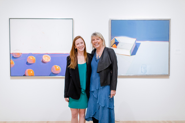 Two women stand with their arms around one another in a museum gallery. They look like mother and daughter and wear blue and black jackets. Behind them are 2 paintings—one has a series of lemons and the depicts a single lemon. 