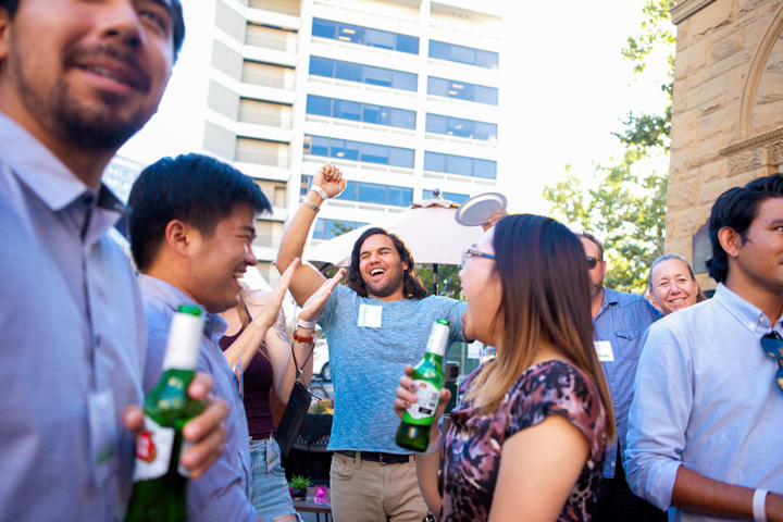 An energetic crowd of people on a patio. A man is raising his fist in the air out of excitement with a huge grin. A man and woman are looking towards each other smiling.