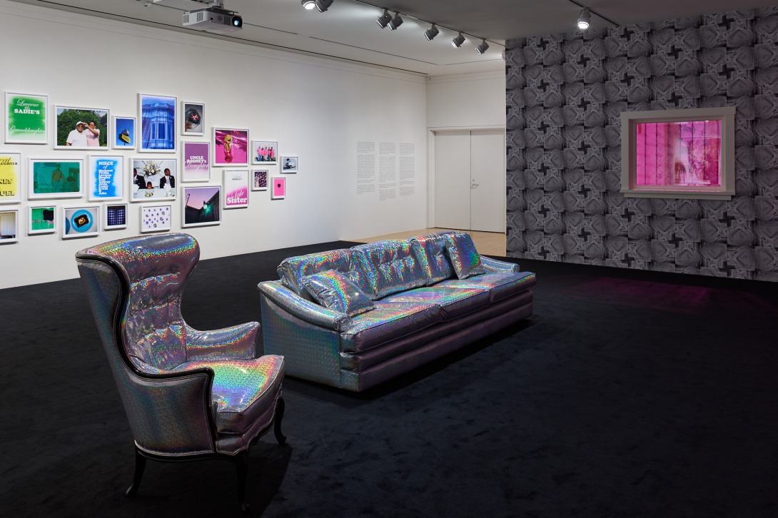 A shiny silver couch and armchair face a wall that is not pictured. A window with bars on it is cut into a wall that has a geometric black and grey wallpaper. Outside the window is a pink glow. Another wall has dozens of colorful framed photos. 