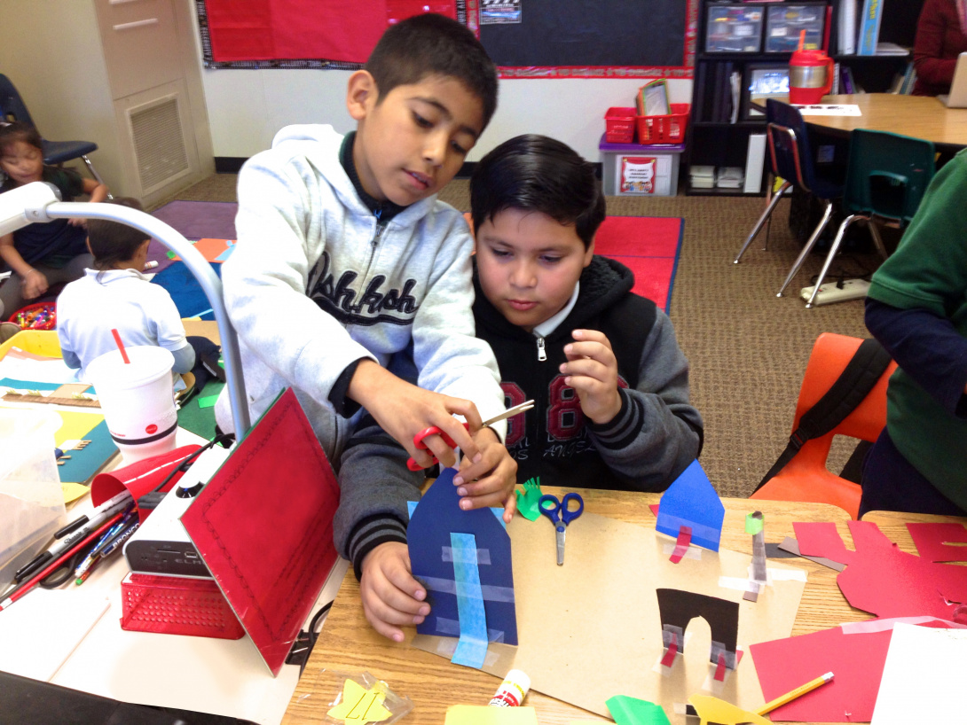 Two young children collaborate on an art project in a classroom. One holds construction paper while the other adheres an object to it. They are creating a cityscape with buildings, houses, and grass by using tape, scissors, and construction paper. 